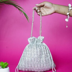 Peut inclure: Un sac à cordon argenté orné de perles et d'un motif en losanges. Le sac est doté de poignées perlées et de pompons décoratifs. Le fond est rose vif, et une petite plante en pot est visible en bas à gauche.