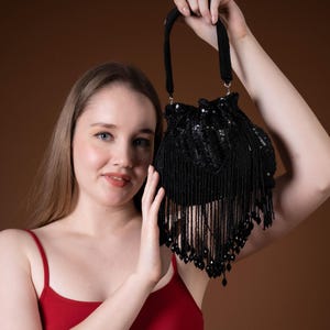 May include: A black beaded handbag with a drawstring closure and long fringe. The bag is held up by a black handle. The bag is decorated with sequins and beads. The background is a solid brown colour.