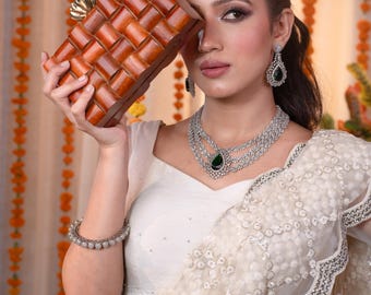 May include: A brown wooden clutch with a woven design and a gold clasp. The image also features a multi-strand silver necklace with a green gemstone, matching earrings, and a bracelet. The model is wearing a white top and a cream-colored, embellished shawl.