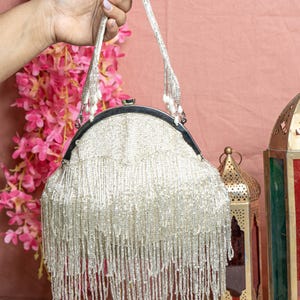 May include: A beaded handbag with a silver frame and a long fringe. The handle is also beaded and has pearl-like accents. The bag is held up against a pink background with pink flowers and decorative lanterns.