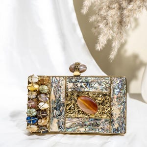 May include: A rectangular clutch with a gold frame and a mix of materials. The front features abalone shell, a large oval stone, and a section with gold filigree. The left side is adorned with a row of colorful, rounded stones.