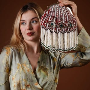 May include: A red and white beaded handbag with pearl fringe. The bag features a decorative design with green accents and a silver clasp. The bag is held up against a brown background.