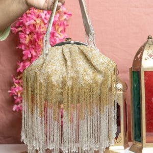 Pode incluir: Uma bolsa com contas com um corpo tecido dourado e creme e um fecho prateado. A bolsa apresenta longas franjas de contas penduradas em ouro e contas transparentes. A alça é creme com contas prateadas. A bolsa é segurada por uma mão.