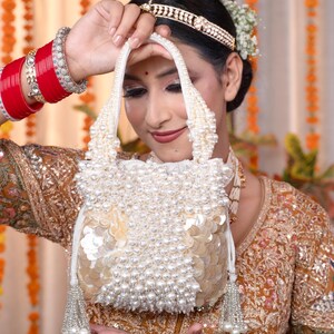 May include: A white beaded handbag with a pearl handle and a sequined base. The bag is held up by a person wearing a gold-colored embroidered outfit and red bangles. The bag has a tassel on each side.