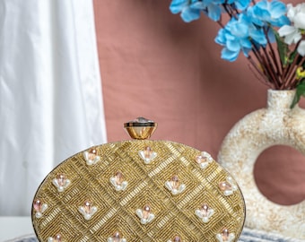 May include: Gold beaded clutch with a diamond pattern, accented with pearls and pink gems. The oval-shaped evening bag has a gold clasp and is set on a blue and white circular mat. A vase of blue flowers is in the background.