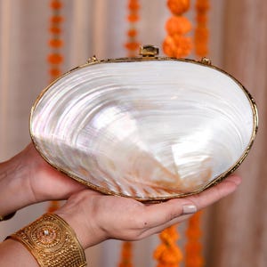 Puede incluir: Un bolso de mano ovalado blanco nacarado con un marco y cierre dorados. El bolso de mano se sostiene en manos abiertas, con una pulsera de brazalete dorada visible en la muñeca. El fondo presenta decoraciones florales naranjas y la marca "VELLESO".
