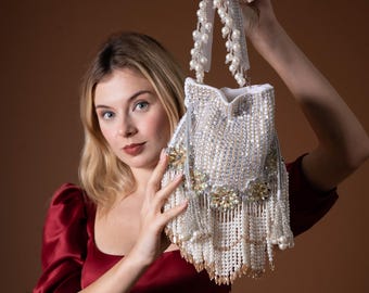 Puede incluir: Un bolso de mano blanco con cuentas, con asa y flecos de perlas. El bolso está adornado con lentejuelas plateadas y detalles dorados. El bolso se sostiene sobre un fondo marrón.