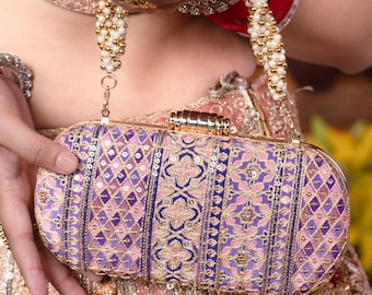 May include: A decorative, oval-shaped clutch in shades of purple and pink, with intricate embroidered patterns and gold accents. The clutch has a gold clasp and a beaded strap. The overall design is ornate and elegant.