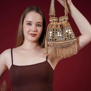 Puede incluir: Un bolso con cordón dorado con diseño de espejo y flecos colgantes. El bolso se sostiene sobre un fondo rojo oscuro. El asa del bolso también está adornada con cuentas.