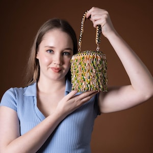 Puede incluir: Un bolso de mano con cuentas de forma cilíndrica. El bolso presenta una mezcla de cuentas verdes, amarillas y color melocotón, con un asa estampada en blanco y negro. El bolso lo sostiene una persona que lleva una camisa azul claro con cuello.