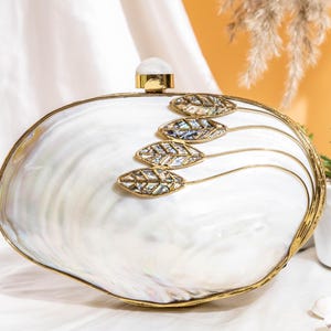 May include: An oval-shaped clutch bag crafted from iridescent white mother-of-pearl with a gold-toned metal frame. The front features four decorative gold-toned leaf motifs inlaid with abalone shell and a round white stone clasp closure.