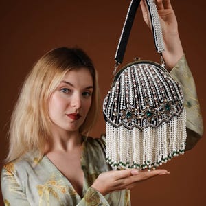May include: An ornate, beaded handbag with a black velvet base and a pearl-adorned handle. The bag features white and pink beads, green gemstones, and a long fringe of white beads. The bag is held up by a person.