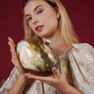 Könnte beinhalten: Ovale Clutch mit natürlicher Horn-Oberfläche in Braun-, Beige- und Cremetönen. Die Clutch hat einen goldfarbenen Rahmen und einen Kettenriemen. Ein weißer Stein verziert den Verschluss. Der Hintergrund ist tiefrot.