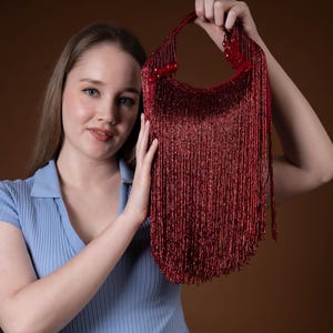 Puede incluir: Un bolso de mano rojo con cuentas y flecos largos. El bolso tiene forma redondeada y un asa hecha con las mismas cuentas rojas. Los flecos cuelgan de la parte inferior del bolso. El bolso es sostenido por una persona.