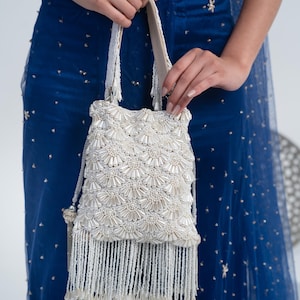 Può includere: Una borsetta bianca con perline con un motivo floreale e lunghe frange di perline. La borsa è tenuta da una persona che indossa un abito blu con abbellimenti dorati. La borsa ha una chiusura con coulisse e una tracolla.