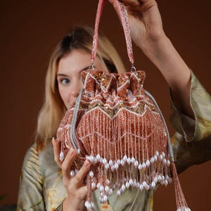 Puede incluir: Un bolso con cordón y cuentas de color melocotón con un asa rosa. El bolso está adornado con lentejuelas doradas y cuentas blancas con forma de perla que cuelgan de la parte inferior. El bolso está sostenido por una persona.