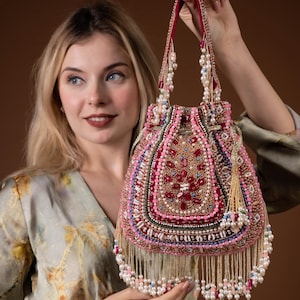 Puede incluir: Un bolso de mano rosa con cuentas, con un asa roja y una borla. El bolso está adornado con cuentas de varios tamaños en tonos rosa, rojo y dorado. La parte inferior del bolso presenta una franja de cuentas blancas. El bolso es sostenido por una persona.