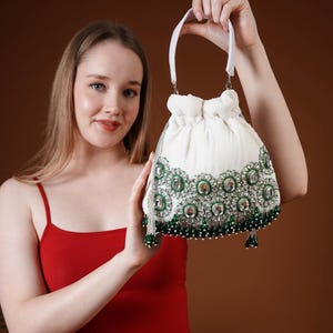 May include: White drawstring bag with a decorative floral pattern in green and silver. The bag has a white handle and is embellished with green beads and silver tassels. The bag is held up by a person wearing a red tank top.