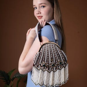 May include: A burgundy beaded handbag with a gold clasp and pearl-like accents. The bag features a decorative design with green gemstones and white fringe. The handle is also adorned with pearls.