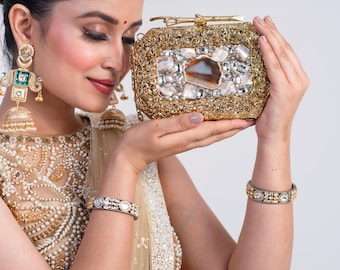 May include: A gold-coloured, rectangular clutch with an ornate, textured design. The clutch features a large, central stone and is embellished with smaller stones. The image also shows a matching bracelet and earrings.