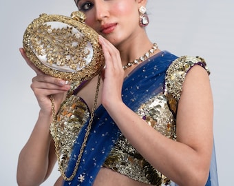 May include: Gold-toned oval clutch with intricate floral detailing and a pearl-like center. The clutch is held by a person wearing a blue and gold embellished outfit, with a matching necklace and earrings. A gold chain strap is attached to the clutch.