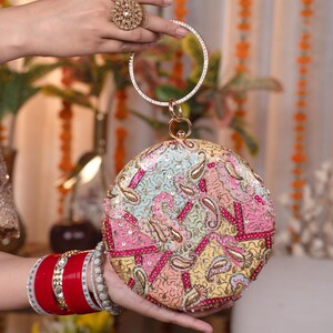Op de afbeelding: Een ronde, veelkleurige clutch met een gouden handvat en ingewikkelde paisley-borduurwerk. De clutch heeft een mix van pastelkleuren, pailletten en gouden accenten. Een grote gouden ring is zichtbaar op een hand die het handvat vasthoudt.