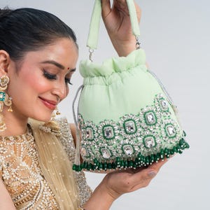 May include: A light green drawstring handbag with silver accents and a beaded fringe. The bag is embellished with large, clear, square-cut rhinestones and smaller green beads. The handle is a matching light green color.