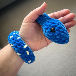 May include: A handmade blue crochet snake toy with a black eye and a matching bracelet. The snake's body is a vibrant blue, with a textured, plush appearance. The bracelet features white accents. The toy is designed to be held.