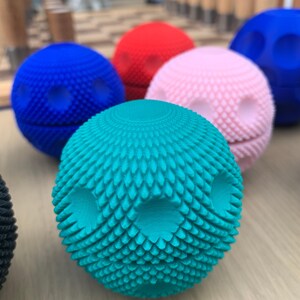 Puede incluir: Una colección de esferas texturizadas y coloridas en tonos turquesa, azul, rojo y rosa. Cada esfera presenta un patrón único y dos hendiduras con forma de ojo. Las esferas están dispuestas sobre una superficie clara, creando una exhibición vibrante. Medidas en cm.
