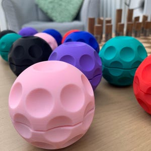 May include: A collection of colorful, spherical objects with a dimpled surface. The spheres are in various colors, including pink, black, purple, blue, and green. The objects are arranged on a light-colored wooden surface.