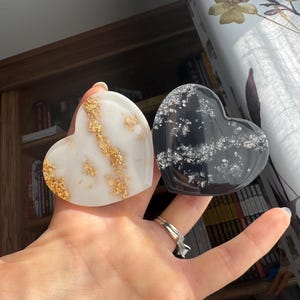 Op de afbeelding: Twee hartvormige harsdecoraties. Eén is wit met goudbladaccenten, de andere is zwart met zilverbladaccenten. De harten worden voor een vage achtergrond gehouden.