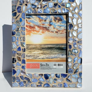 May include: Rectangular photo frame with a mosaic border of blue and iridescent shell pieces. The frame holds a picture of a sunset over the ocean. The frame is 5 inches by 7 inches.