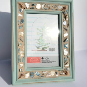 May include: A light blue picture frame with a decorative border made of seashells and sand. The frame is 4 inches by 6 inches (10.16 cm x 15.24 cm). The frame is made by Greentree Gallery.