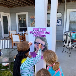 Puede incluir: Un juego de fiesta llamado "Pon las gafas a Ruthie" se muestra en un letrero blanco. Los ni&ntilde;os est&aacute;n jugando, tratando de colocar gafas en la imagen de una ni&ntilde;a. El letrero est&aacute; montado en un pilar blanco, con una casa y muebles de exterior en el fondo.