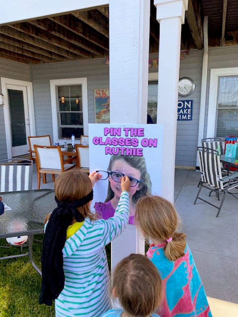 Puede incluir: Un juego de fiesta llamado "Pon las gafas a Ruthie" se muestra en un letrero blanco. Los ni&ntilde;os est&aacute;n jugando, tratando de colocar gafas en la imagen de una ni&ntilde;a. El letrero est&aacute; montado en un pilar blanco, con una casa y muebles de exterior en el fondo.