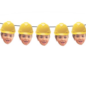 Puede incluir: Una guirnalda decorativa con cinco caras sonrientes que llevan cascos amarillos de construcción. Las caras están ensartadas en una cuerda negra sobre un fondo blanco.