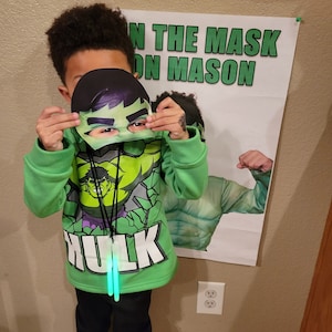Puede incluir: Un ni&ntilde;o con una sudadera con capucha verde con tem&aacute;tica de Hulk y pantalones negros, sosteniendo una m&aacute;scara de Hulk. La sudadera tiene un gr&aacute;fico de Hulk y la palabra "Hulk". Un p&oacute;ster con el texto "ON THE MASK ON MASON" est&aacute; en el fondo.
