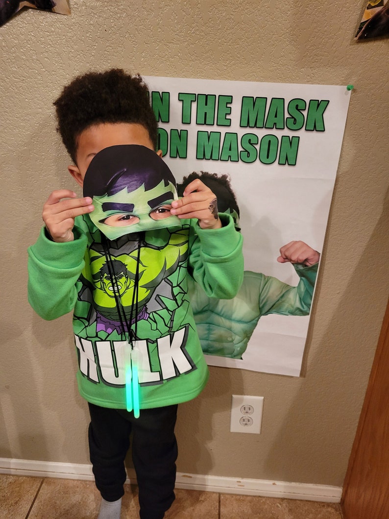 Puede incluir: Un ni&ntilde;o con una sudadera con capucha verde con tem&aacute;tica de Hulk y pantalones negros, sosteniendo una m&aacute;scara de Hulk. La sudadera tiene un gr&aacute;fico de Hulk y la palabra "Hulk". Un p&oacute;ster con el texto "ON THE MASK ON MASON" est&aacute; en el fondo.