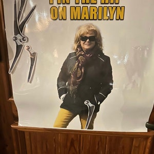 Puede incluir: Un cartel con el texto "PIN THE HIP ON MARILYN" en letras doradas. El cartel presenta a una mujer con gafas de sol, una chaqueta negra y pantalones amarillos. Diagramas de articulaciones de la cadera se colocan alrededor de la mujer.
