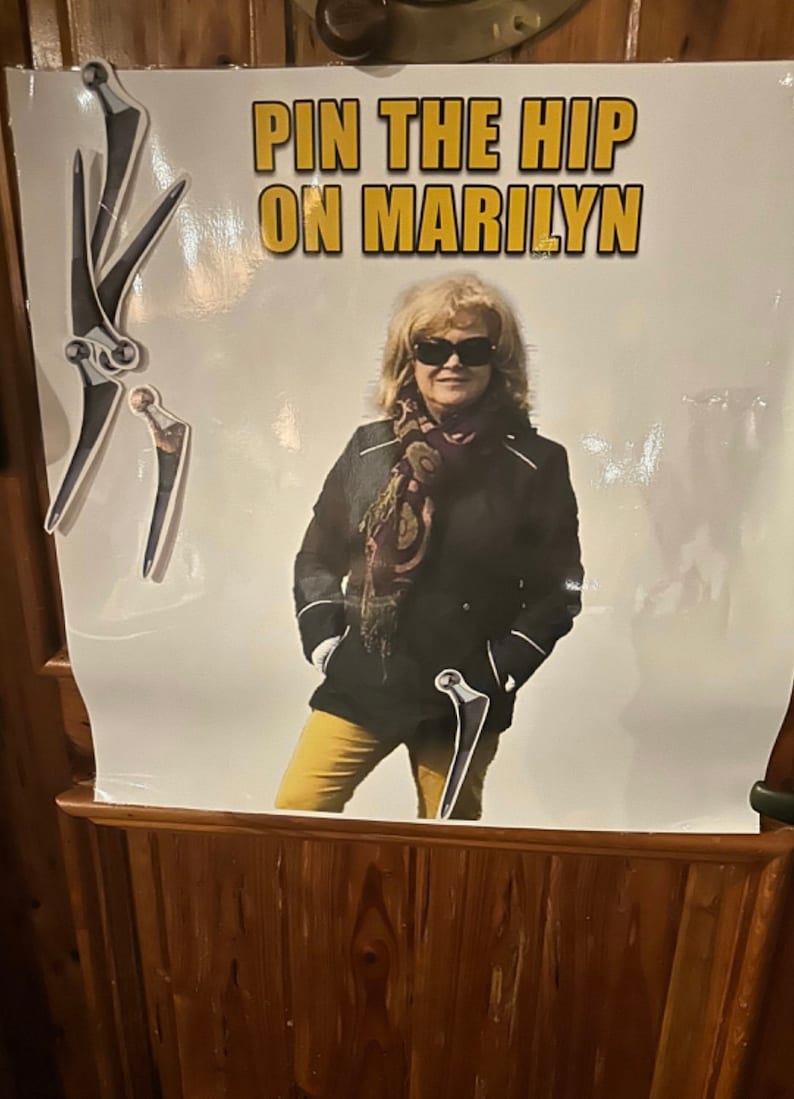 Puede incluir: Un cartel con el texto "PIN THE HIP ON MARILYN" en letras doradas. El cartel presenta a una mujer con gafas de sol, una chaqueta negra y pantalones amarillos. Diagramas de articulaciones de la cadera se colocan alrededor de la mujer.
