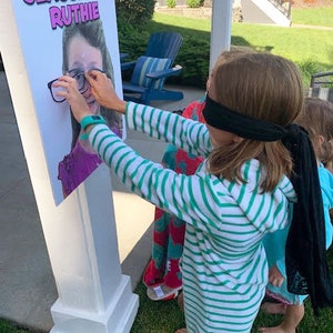 Puede incluir: Un soporte de madera blanco con un p&oacute;ster que dice "Pin the Glasses on Ruthie". Un ni&ntilde;o con un vestido con capucha a rayas verdes y blancas y una venda negra intenta poner gafas en el p&oacute;ster. Un divertido juego de fiesta.