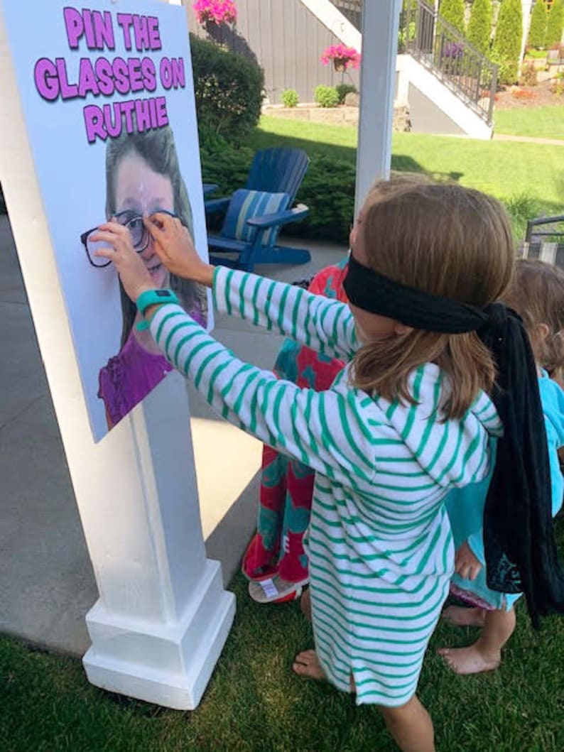 Puede incluir: Un soporte de madera blanco con un p&oacute;ster que dice "Pin the Glasses on Ruthie". Un ni&ntilde;o con un vestido con capucha a rayas verdes y blancas y una venda negra intenta poner gafas en el p&oacute;ster. Un divertido juego de fiesta.