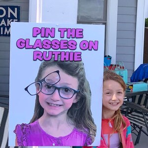 Puede incluir: Un letrero con el texto "PIN THE GLASSES ON RUTHIE" presenta a una ni&ntilde;a sonriente con gafas. El letrero lo sostiene otra ni&ntilde;a que lleva una chaqueta rosa y naranja.