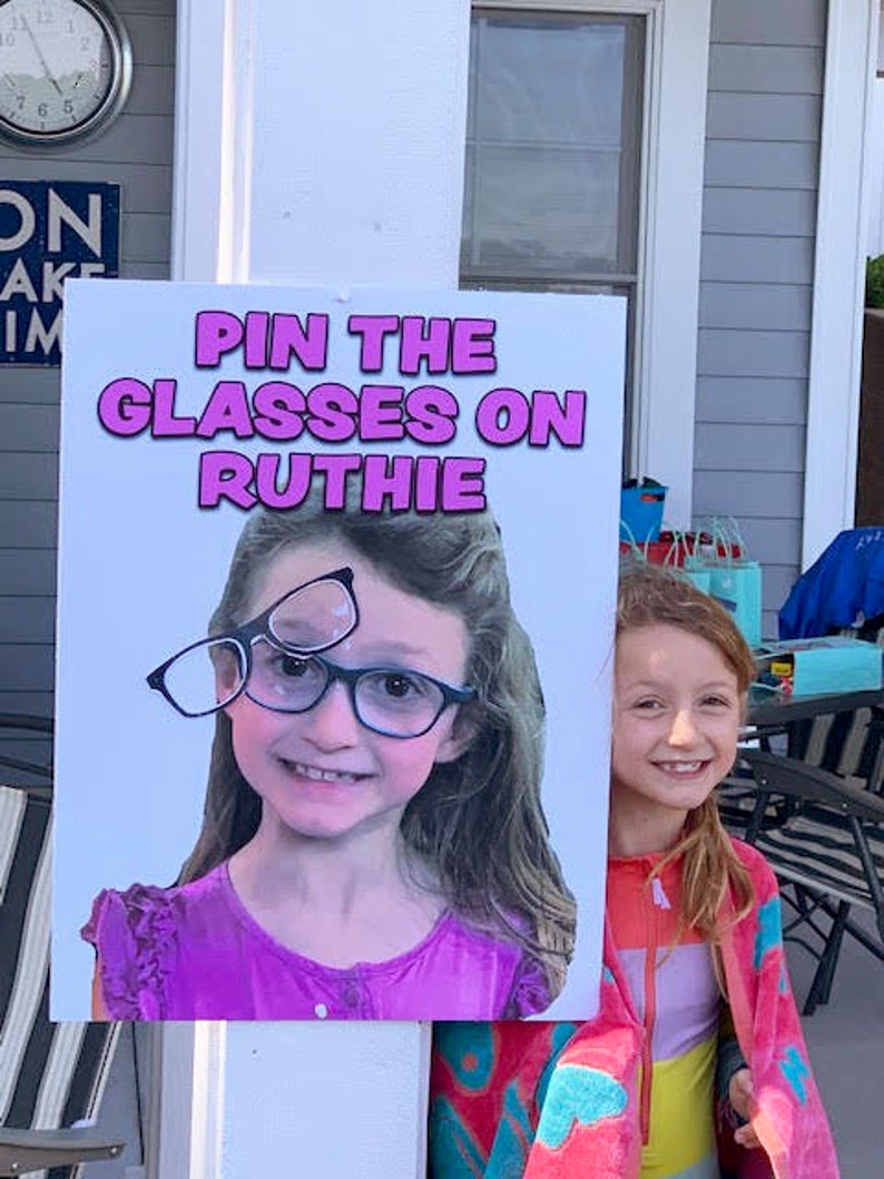Puede incluir: Un letrero con el texto "PIN THE GLASSES ON RUTHIE" presenta a una ni&ntilde;a sonriente con gafas. El letrero lo sostiene otra ni&ntilde;a que lleva una chaqueta rosa y naranja.
