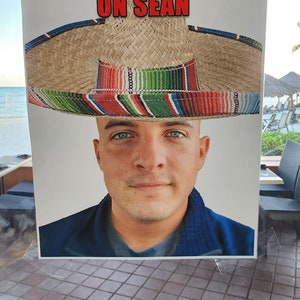 Puede incluir: Un letrero impreso con el texto "PIN THE MUSTACHE ON SEAN" en letras rojas. El letrero presenta a un hombre con un sombrero. El sombrero es de paja con una banda a rayas coloridas.