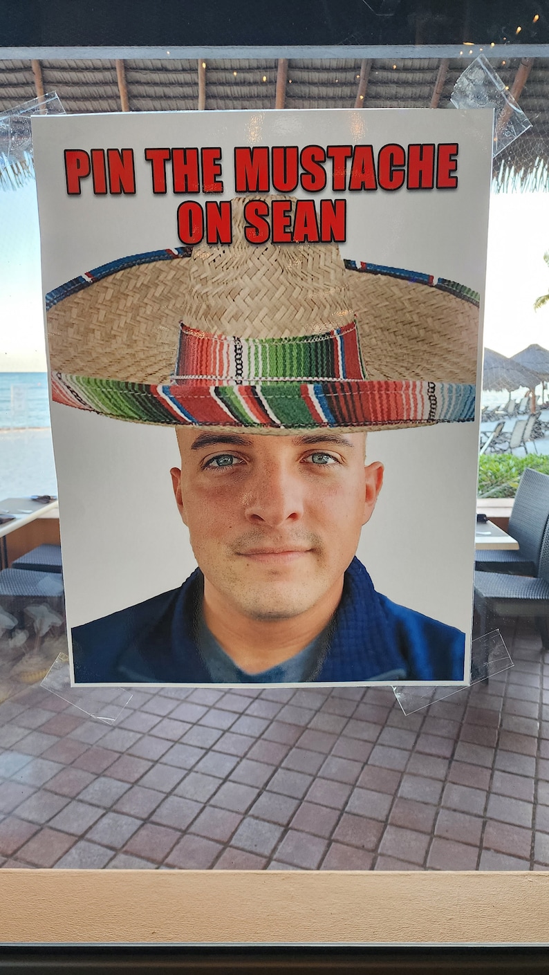 Puede incluir: Un letrero impreso con el texto "PIN THE MUSTACHE ON SEAN" en letras rojas. El letrero presenta a un hombre con un sombrero. El sombrero es de paja con una banda a rayas coloridas.