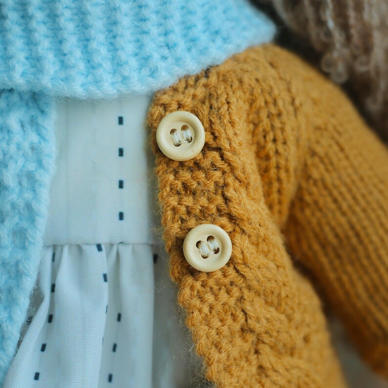 K&ouml;nnte beinhalten: Nahaufnahme eines handgefertigten Puppenoutfits. Das Outfit umfasst einen hellblauen Strickschal, eine senfgelbe Strickjacke mit zwei Holzkn&ouml;pfen und ein wei&szlig;es Kleid mit kleinem blauen Tupfenmuster.