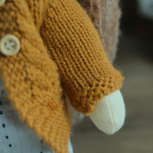 K&ouml;nnte beinhalten: Nahaufnahme eines handgefertigten Puppenarms und eines Teils ihres K&ouml;rpers. Die Puppe tr&auml;gt eine senfgelbe Strickjacke mit Holzkn&ouml;pfen. Der Arm der Puppe ist aus cremefarbenem Stoff.