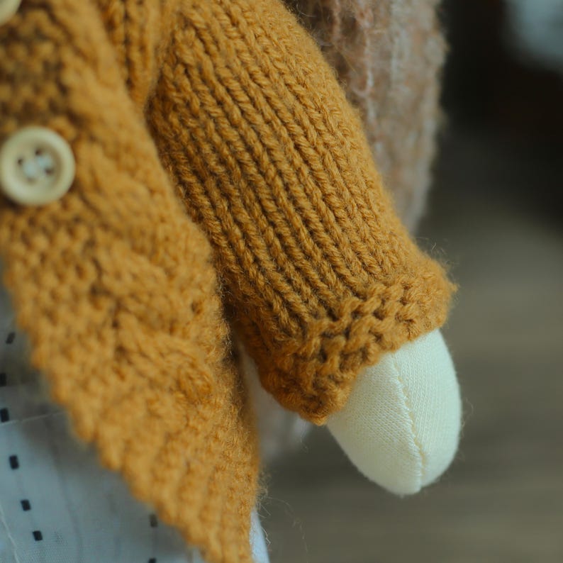 K&ouml;nnte beinhalten: Nahaufnahme eines handgefertigten Puppenarms und eines Teils ihres K&ouml;rpers. Die Puppe tr&auml;gt eine senfgelbe Strickjacke mit Holzkn&ouml;pfen. Der Arm der Puppe ist aus cremefarbenem Stoff.