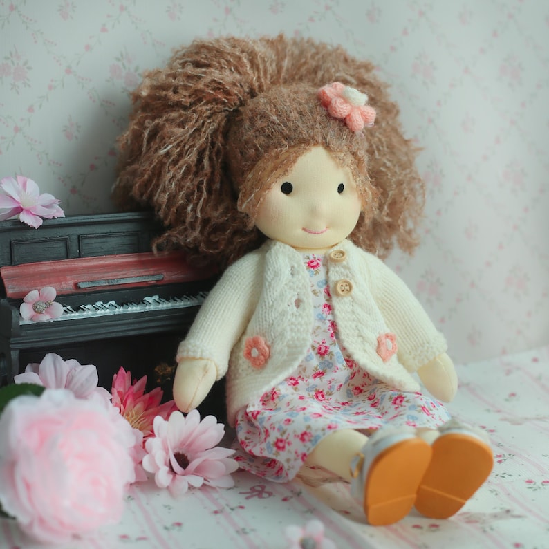 May include: A handmade doll with brown curly hair and a pink flower hair accessory. The doll wears a floral print dress, a cream-colored cardigan with flower accents, and silver shoes with orange soles. The doll is sitting near a miniature piano.